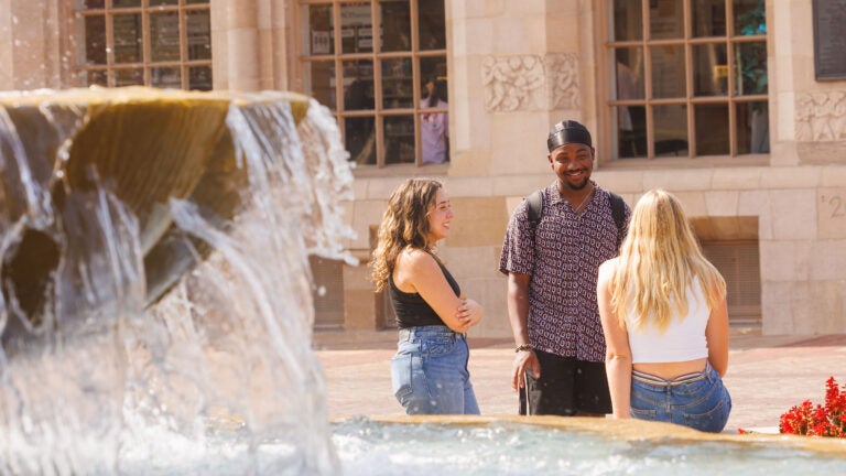 Three students talk in front of a fountain on a sunny day.